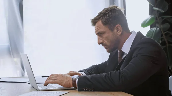 Vista lateral del hombre de negocios en traje escribiendo en el ordenador portátil en la oficina - foto de stock