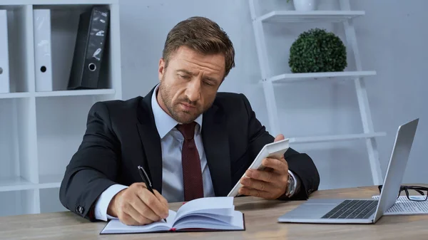 Homem de negócios com calculadora escrevendo em notebook perto de laptop no local de trabalho — Fotografia de Stock