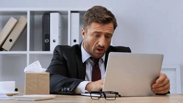 Hombre se siente mal mientras trabaja en el portátil cerca de servilletas de papel en el escritorio de la oficina - foto de stock