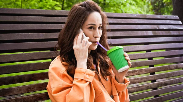 Femme boire du café pour passer à travers la paille et parler sur smartphone dans le parc — Photo de stock