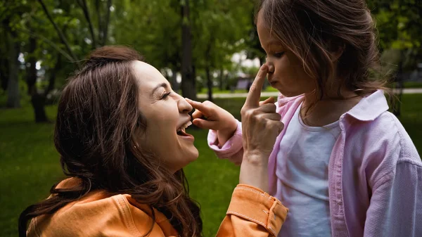 Vue latérale de la mère étonnée touchant le nez de la fille mignonne dans le parc vert — Photo de stock