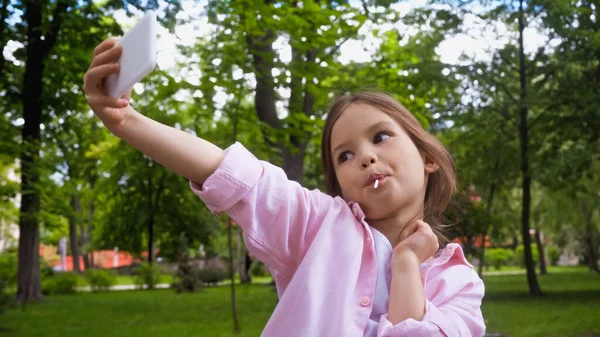 Kid with lollipop on stick taking selfie on smartphone in park — Stock Photo