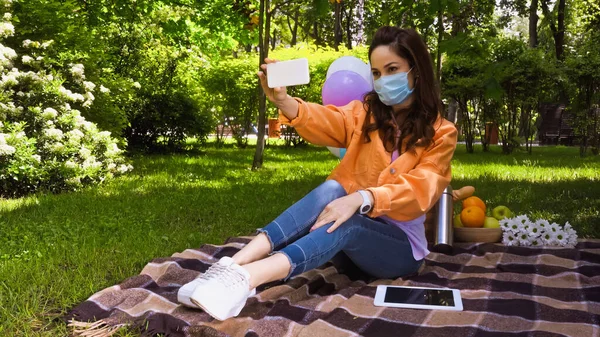 Woman in medical mask taking selfie on smartphone near digital tablet in park — Stock Photo