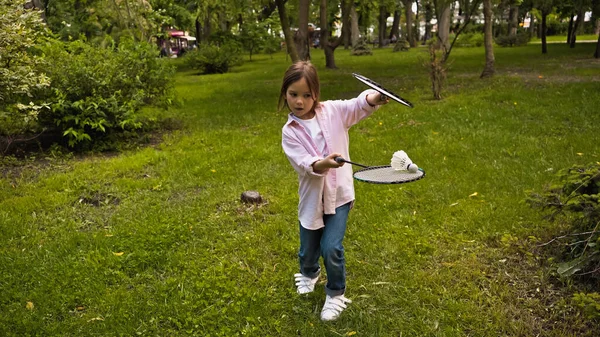 Funny kid in casual clothing playing badminton on green grass in park — Foto stock