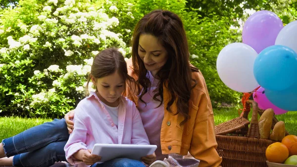 Glückliche Mutter und Tochter schauen auf digitales Tablet, während sie im Park neben Luftballons sitzen — Stockfoto