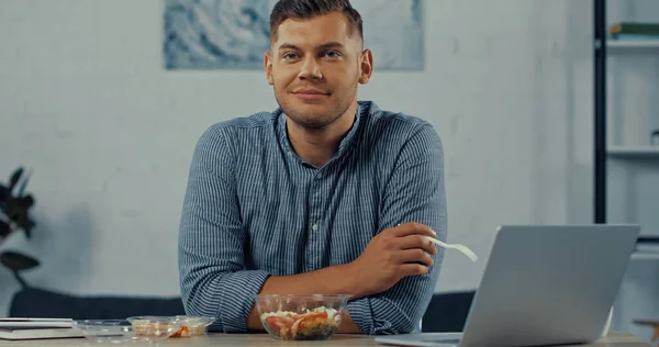 Homme heureux tenant fourchette en plastique près de la salade dans le récipient et ordinateur portable sur le bureau — Photo de stock