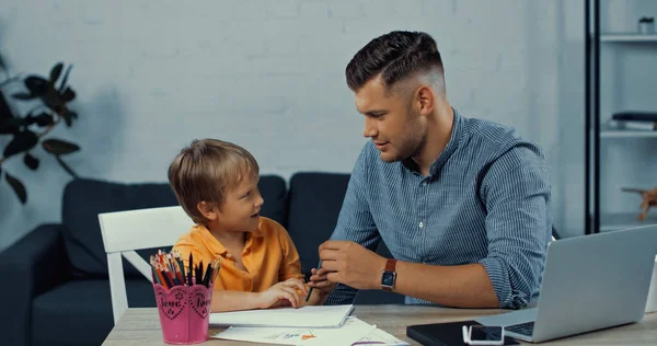 Vater hält Bleistift in der Hand und schaut Sohn neben Spielzeug auf dem Tisch an — Stockfoto