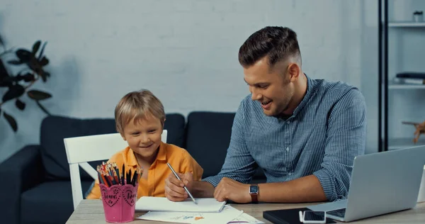 Щасливий батько малює на папері біля веселого сина з гаджетами на столі. — стокове фото