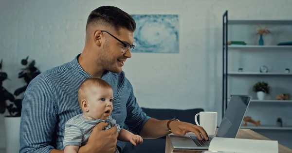 Cheerful father in glasses using laptop while infant boy and working from home — Stockfoto
