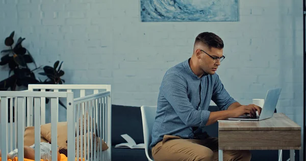 Freelancer en gafas usando laptop mientras está sentado cerca de una cuna con hijo pequeño - foto de stock