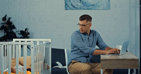 Stressed freelancer in glasses using laptop while sitting near baby crib with infant son — Stock Photo