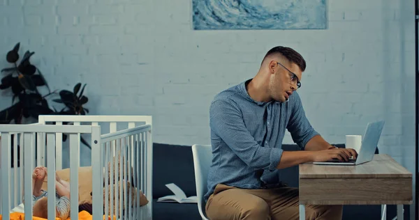 Freelancer in glasses talking on smartphone and using laptop while sitting near baby crib with infant son — Stock Photo