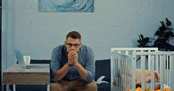 Worried father in eyeglasses sitting near baby crib with infant son and desk with gadgets — Foto stock