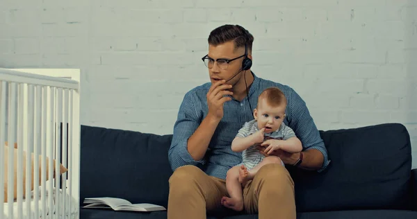 Людина в гарнітурі з мікрофоном тримає немовляти сина під час роботи з дому — стокове фото