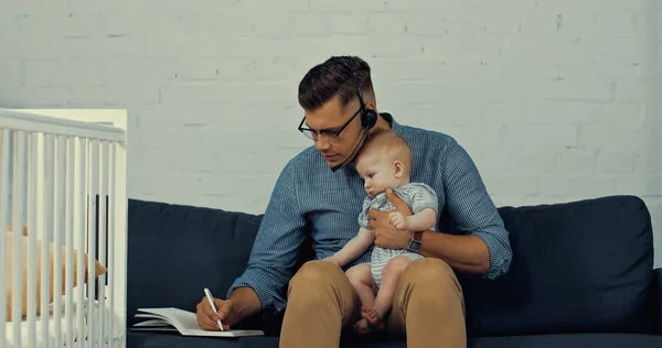 Людина в гарнітурі з мікрофоном тримає немовляти сина під час написання в блокноті і роботи з дому — стокове фото