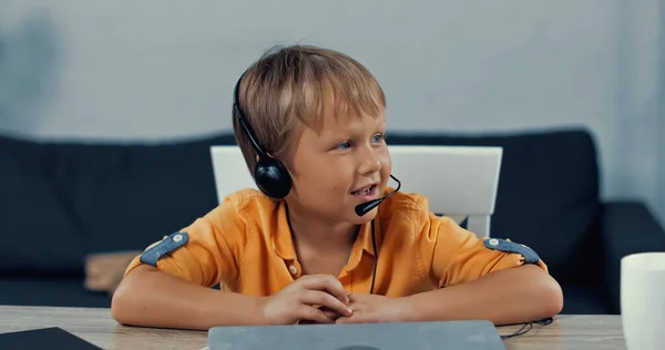 Щасливий хлопчик в гарнітурі з мікрофоном, що розмовляє біля ноутбука на столі — стокове фото