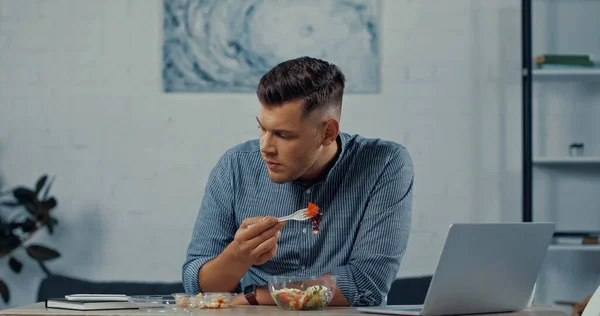 Freelancer holding plastic fork and eating salad near laptop on desk — Stock Photo