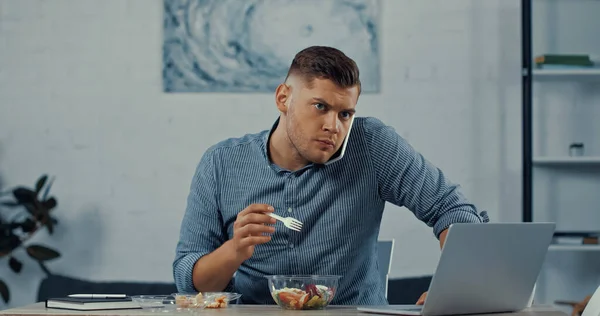 Freelancer holding plastic fork and eating salad while talking on smartphone near laptop on desk — Foto stock