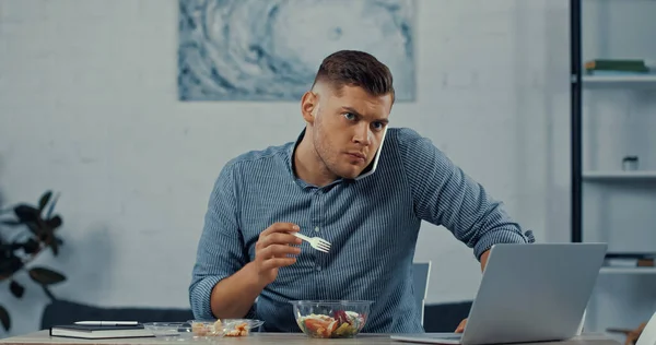 Freelancer holding plastic fork and eating salad while talking on smartphone near laptop on desk — Stock Photo