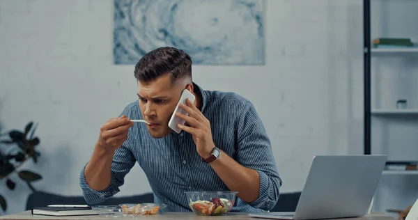 Homem segurando garfo de plástico e comer salada enquanto conversa no smartphone perto do laptop na mesa — Fotografia de Stock