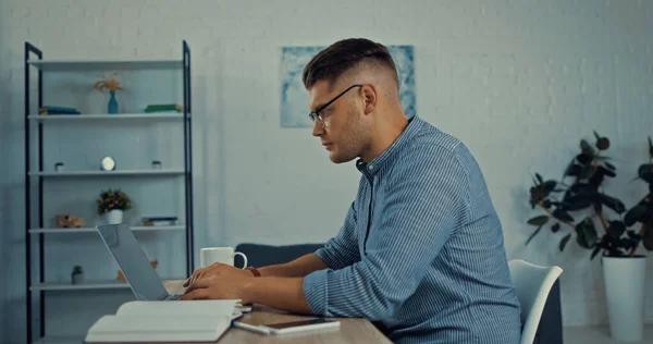 Боковой вид фрилансера в очках, использующего ноутбук рядом со смартфоном на столе во время работы из дома — стоковое фото