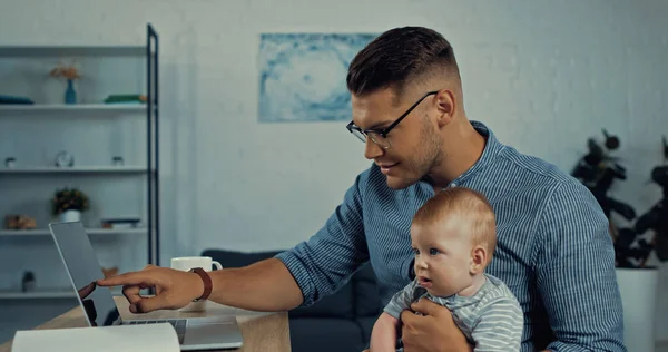 Fröhlicher Freiberufler mit Brille zeigt auf Laptop, während er kleinen Jungen hält — Stockfoto