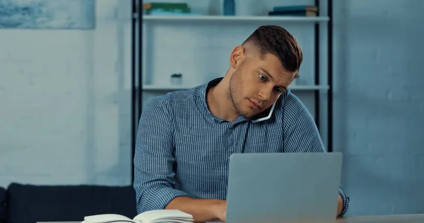 Freelancer ocupado hablando en el teléfono inteligente y el uso de la computadora portátil mientras trabaja desde casa - foto de stock