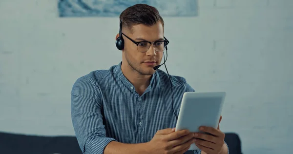 Teletrabajador en auriculares con micrófono que sostiene la tableta digital mientras trabaja desde casa - foto de stock