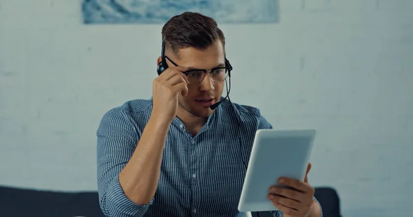 Teleworker in headset with microphone adjusting glasses while holding digital tablet and working from home - foto de stock