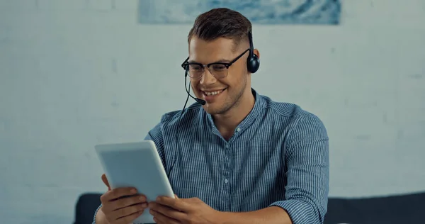 Teletrabajador feliz en auriculares con micrófono que sostiene la tableta digital mientras trabaja desde casa - foto de stock