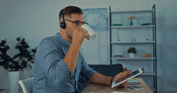 Teletrabajador en vasos bebiendo café mientras sostiene la tableta digital cerca de dispositivos - foto de stock