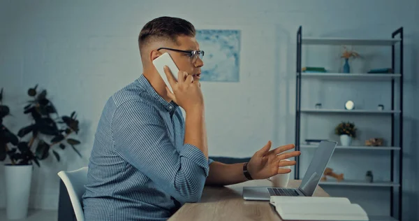 Vue latérale de l'homme en lunettes parlant sur smartphone tout en travaillant à distance à la maison — Photo de stock