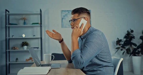 Side view of freelancer in glasses talking on smartphone while working remotely at home — Fotografia de Stock