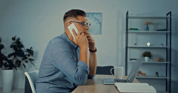 Side view of worried man in glasses talking on smartphone while working remotely at home — Stock Photo