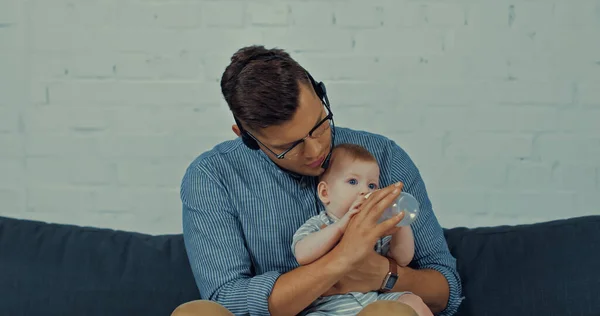 Teleworker in headset with microphone feeding infant boy with milk in baby bottle - foto de stock