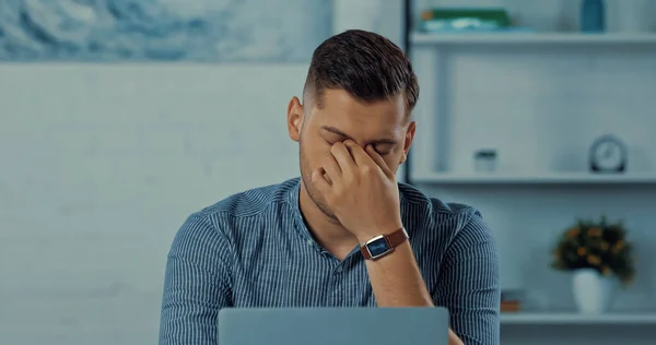 Freelancer cansado tocando los ojos cerca del ordenador portátil mientras trabaja desde casa - foto de stock