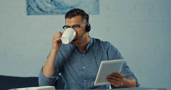 Operator in headset and glasses drinking coffee and using digital tablet — Fotografia de Stock