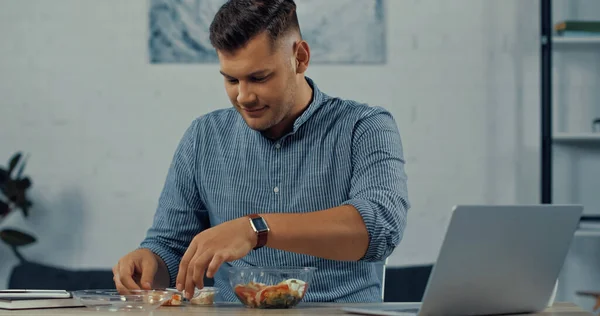 Uomo sorridente guardando insalata in contenitore di plastica vicino al computer portatile sulla scrivania — Foto stock