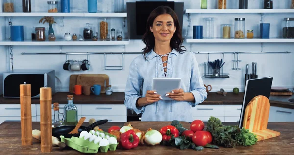 Mujer vegetariana con tableta digital sonriendo a la cámara cerca de ingredientes crudos en la cocina - foto de stock