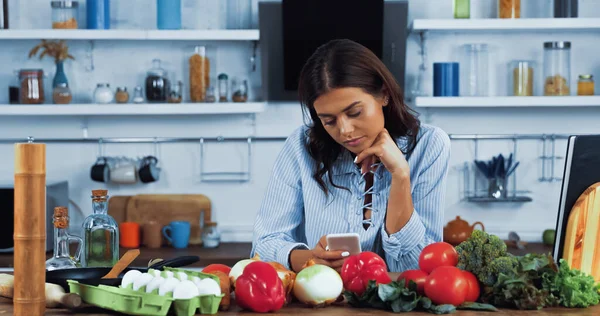 Woman using smartphone near various ingredients and cookbook on kitchen table — Stockfoto