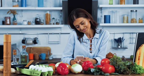 Donna sorridente che legge la ricetta in smartphone vicino a verdure fresche e uova in cucina — Foto stock