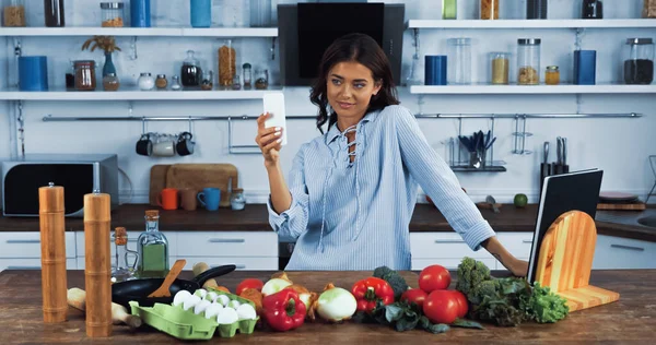 Счастливая женщина делает селфи на смартфоне рядом с сырыми ингредиентами на кухонном столе — стоковое фото