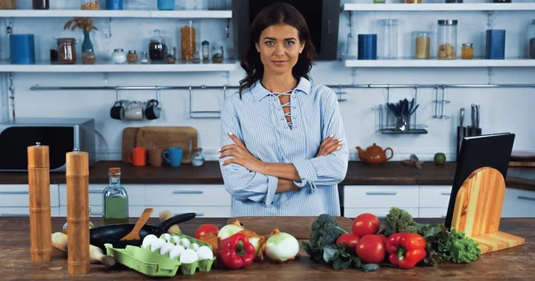 Mulher morena com braços cruzados sorrindo para a câmera perto de diferentes vegetais e ovos — Fotografia de Stock