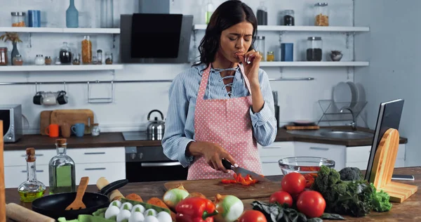 Casalinga con coltello leccare il dito tagliato vicino al peperone tritato e verdure fresche — Foto stock