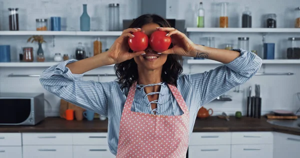 Alegre morena mujer en delantal cubriendo los ojos con tomates rojos maduros - foto de stock