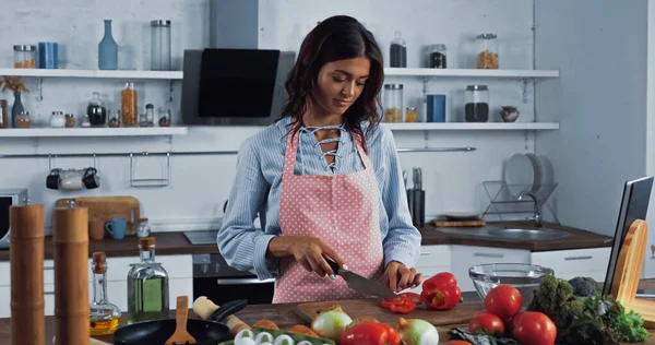 Brunette woman in apron cutting bell pepper while cooking in kitchen — Foto stock