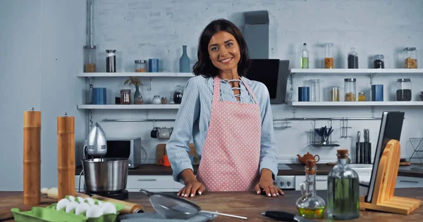 Femme heureuse dans le tablier près du robot culinaire et des ingrédients sur le plan de travail de la cuisine — Photo de stock