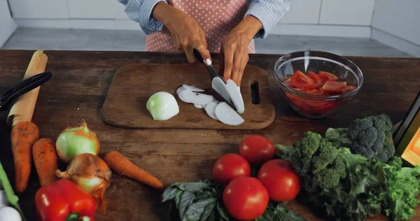 Partial view of housewife cutting fresh onion near raw vegetables and bowl with chopped tomatoes - foto de stock