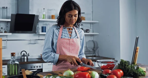 Woman in apron cutting bell pepper near various vegetables on kitchen worktop — Stockfoto