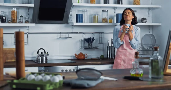 Ama de casa complacida con taza de té cerca de encimera con alimentos y utensilios de cocina en primer plano borroso - foto de stock